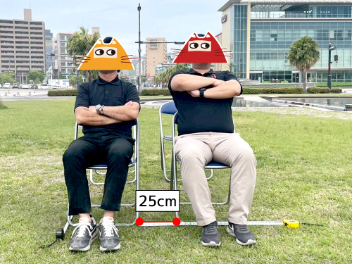 パイプ椅子2脚に着席する男性二人。パイプ椅子の横間隔は25cm。