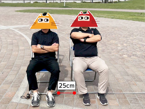 パイプ椅子2脚に着席する男性二人。パイプ椅子の横間隔は25cm。