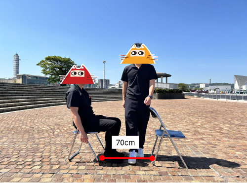 パイプ椅子の前後間隔は70cm。男性Aが着席、男性Bがその前を横切る様子