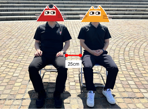 パイプ椅子2脚に着席する男性二人。パイプ椅子の横間隔は25cm。